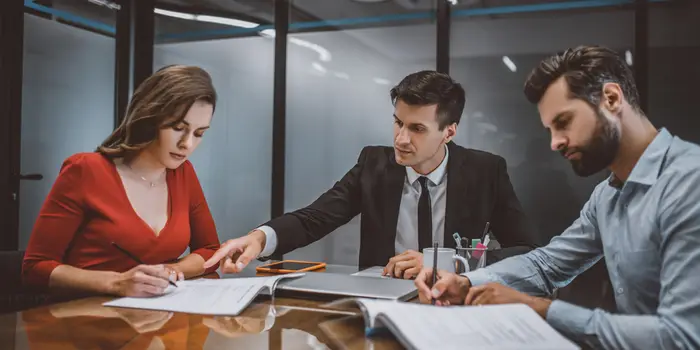 An attractive couple sit on opposite sides of a table separated by a solicitor as they sign legal documents